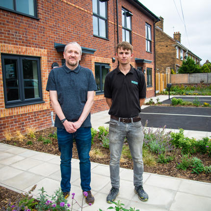 Two people stand on a paved walkway outside a row of newly built red-brick homes. Landscaped flower beds and planting border the path, with modern black-framed windows, parking spaces and neighbouring properties visible in the background.
