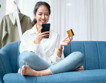 Person sitting on a blue couch, casually dressed in a white shirt and jeans, holding a smartphone in one hand and a credit card in the other, suggesting an online payment or shopping activity. An olive green jacket hangs on a white ladder in the background.