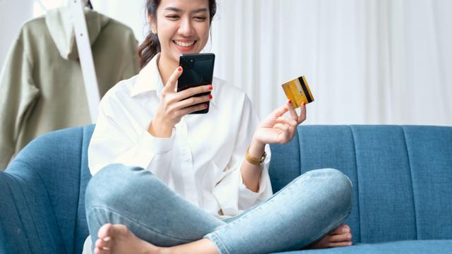 Person sitting on a blue couch, casually dressed in a white shirt and jeans, holding a smartphone in one hand and a credit card in the other, suggesting an online payment or shopping activity. An olive green jacket hangs on a white ladder in the background.