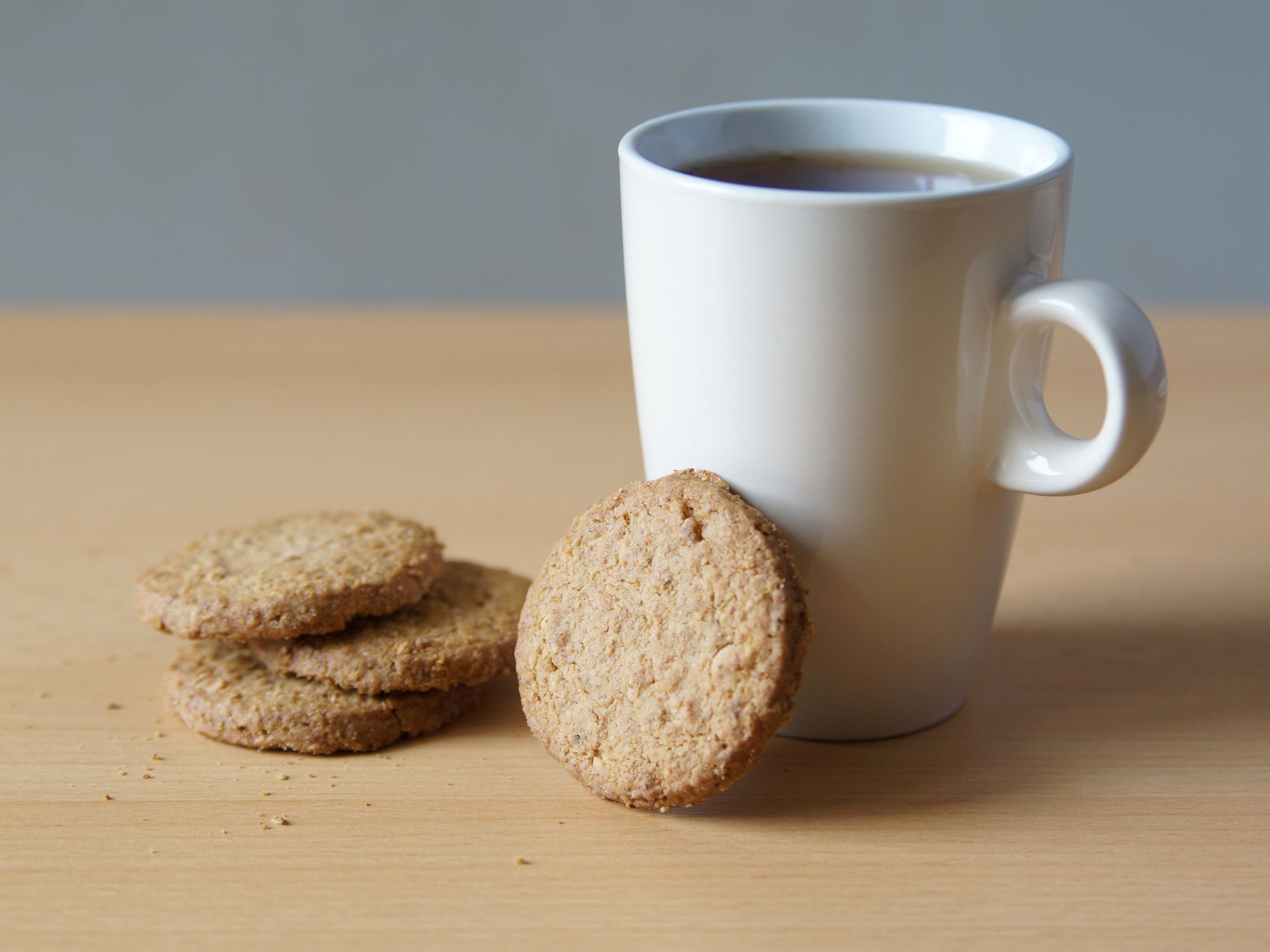 A white mug filled with tea or coffee on a wooden surface, accompanied by four round cookies, suggesting a warm and inviting snack moment.