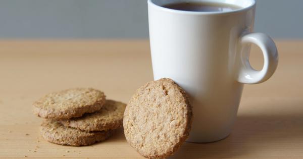 A white mug filled with tea or coffee on a wooden surface, accompanied by four round cookies, suggesting a warm and inviting snack moment.