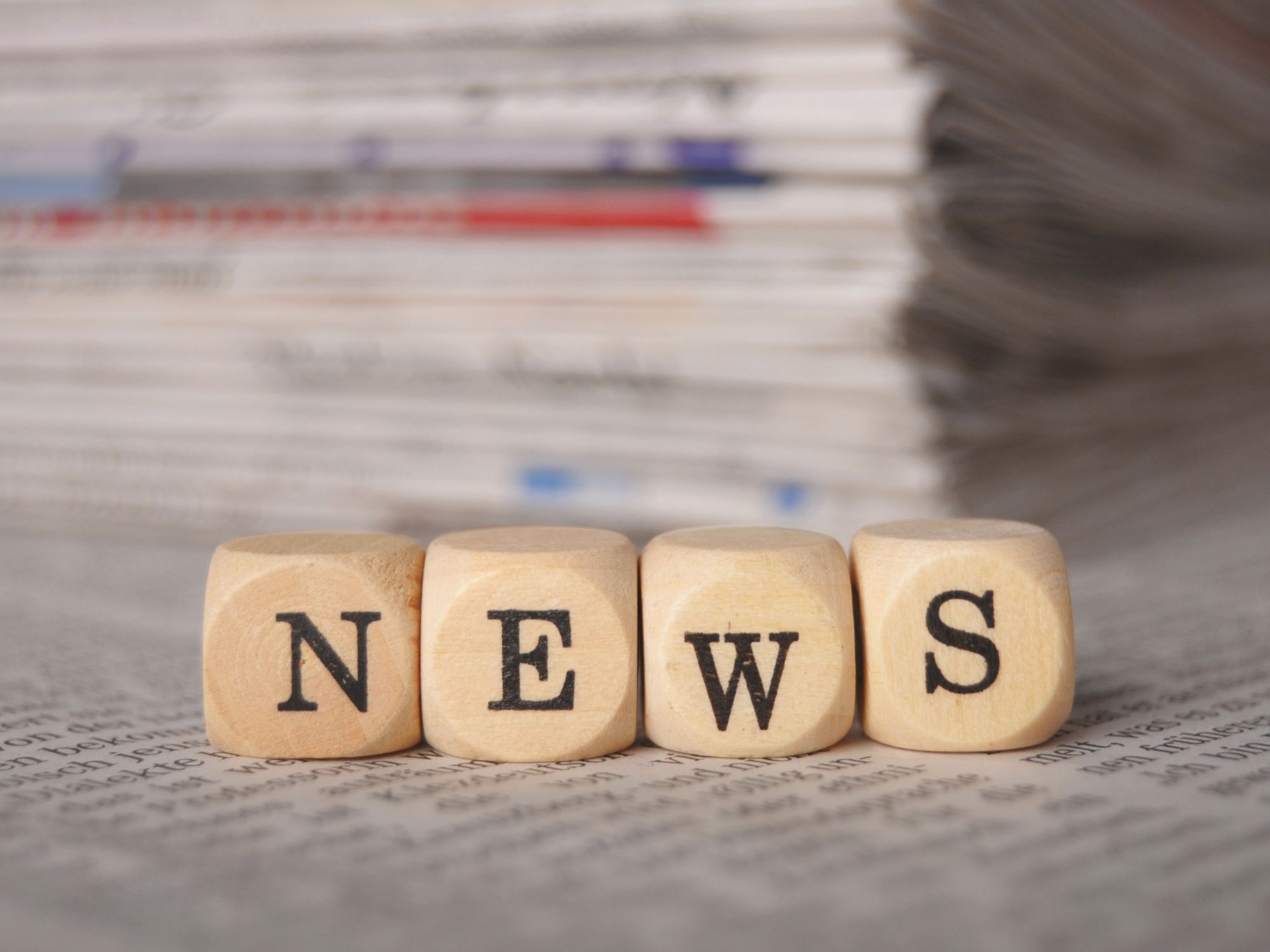 Four wooden blocks spelling “NEWS” placed on top of a newspaper, with a stack of newspapers in the background.