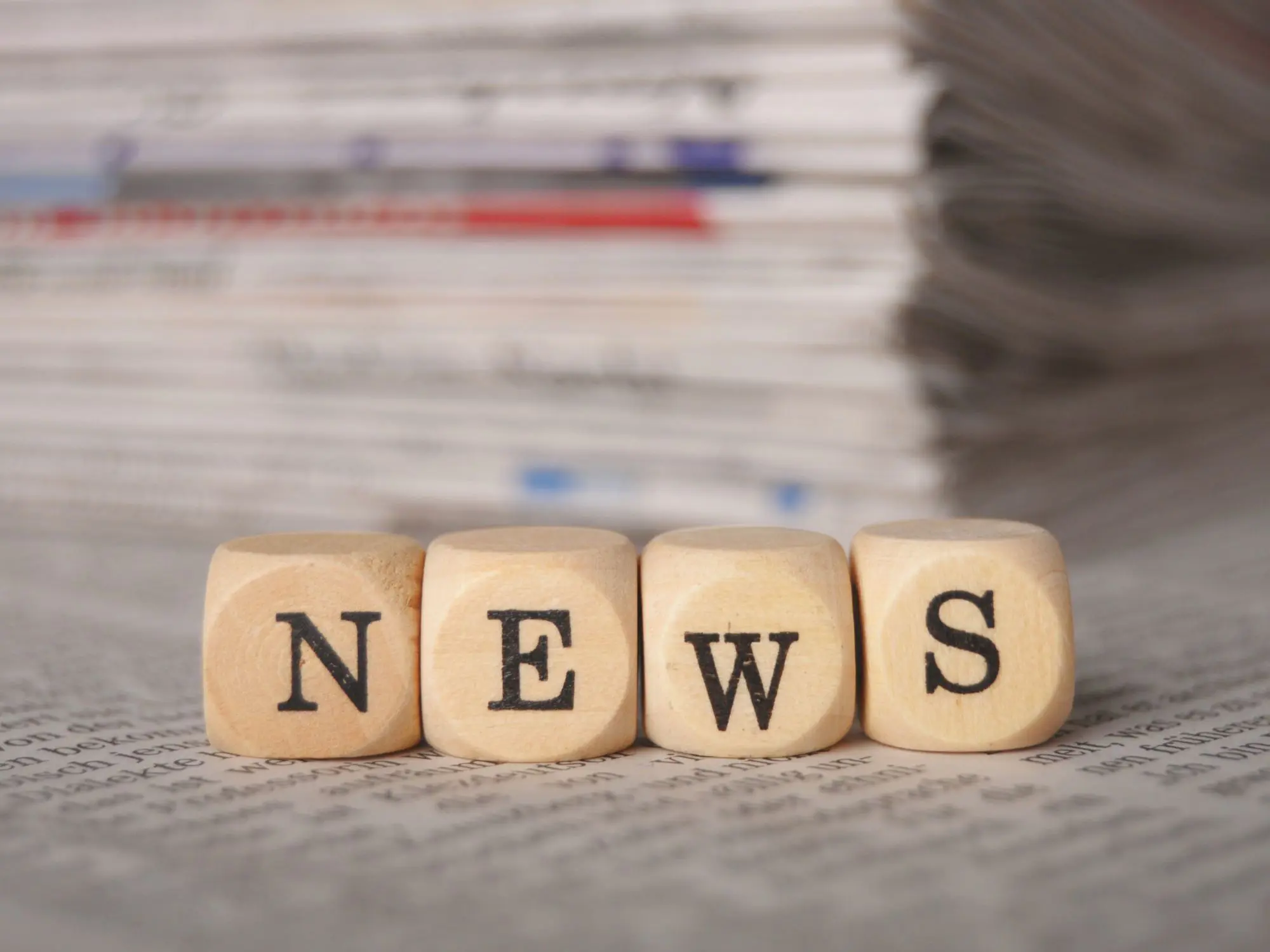 Four wooden blocks spelling “NEWS” placed on top of a newspaper, with a stack of newspapers in the background.
