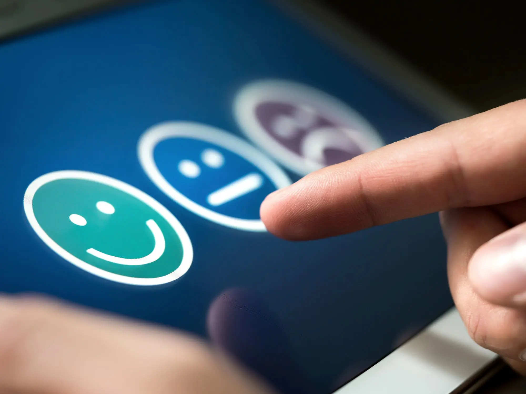 A hand interacting with a touchscreen displaying three emoticons—green smiling face, neutral face, and purple sad face—indicating a positive feedback selection.