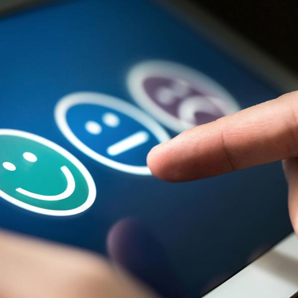 A hand interacting with a touchscreen displaying three emoticons—green smiling face, neutral face, and purple sad face—indicating a positive feedback selection.