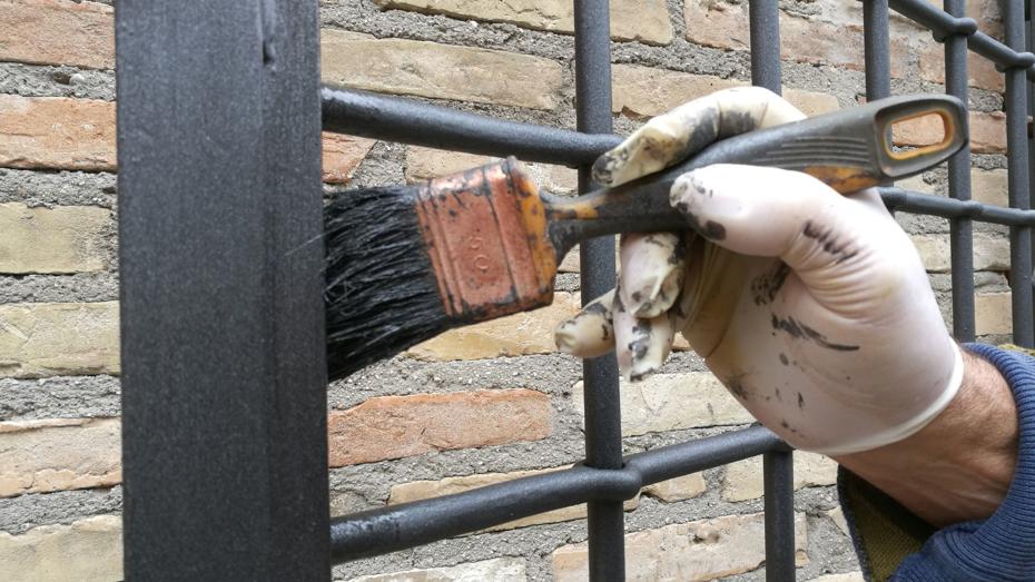 Gloved hand painting a black metal gate in front of a brick wall.