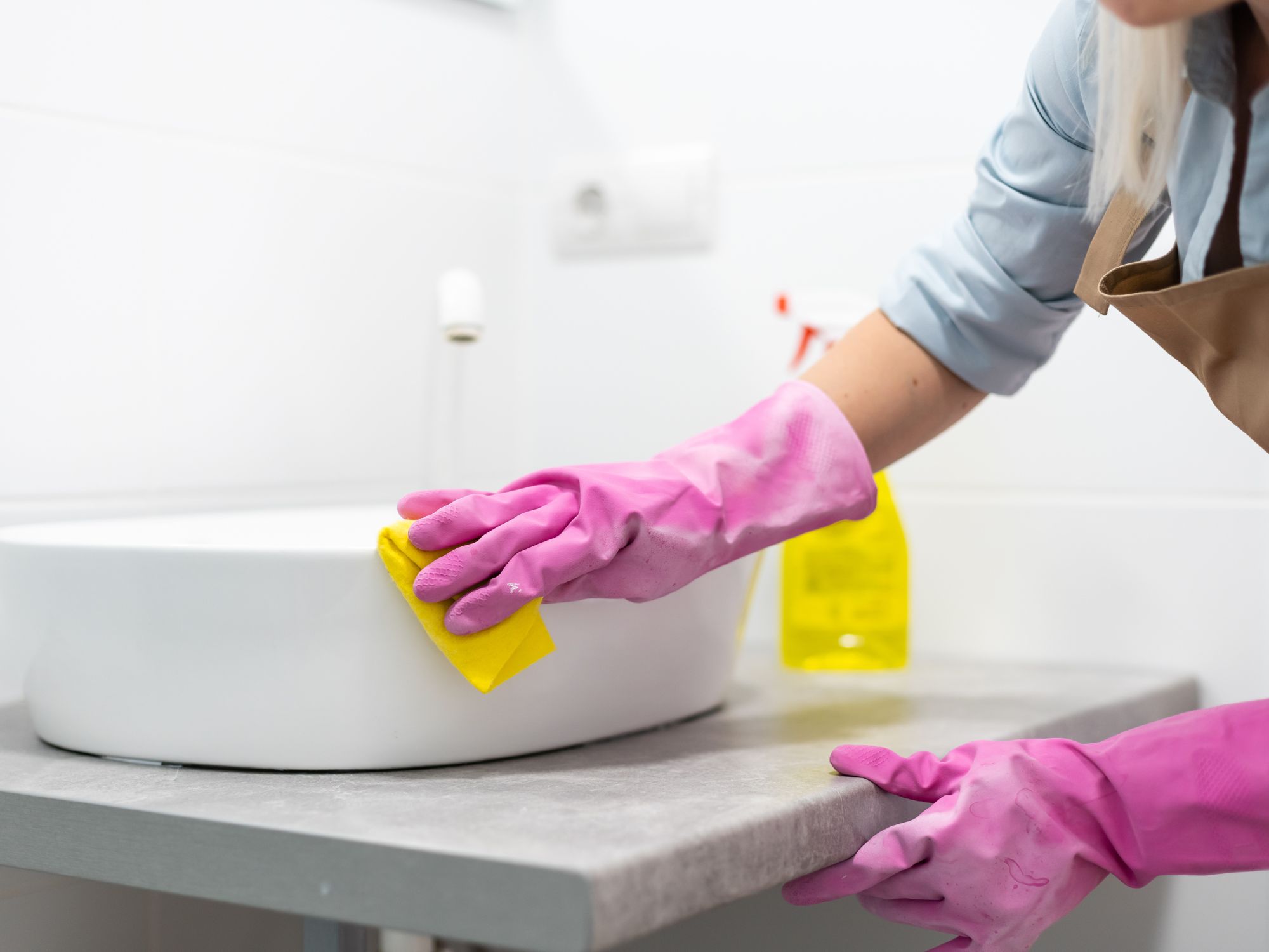 Person wearing pink rubber gloves cleaning a bathroom sink with a yellow sponge; a spray bottle with yellow liquid is visible in the background