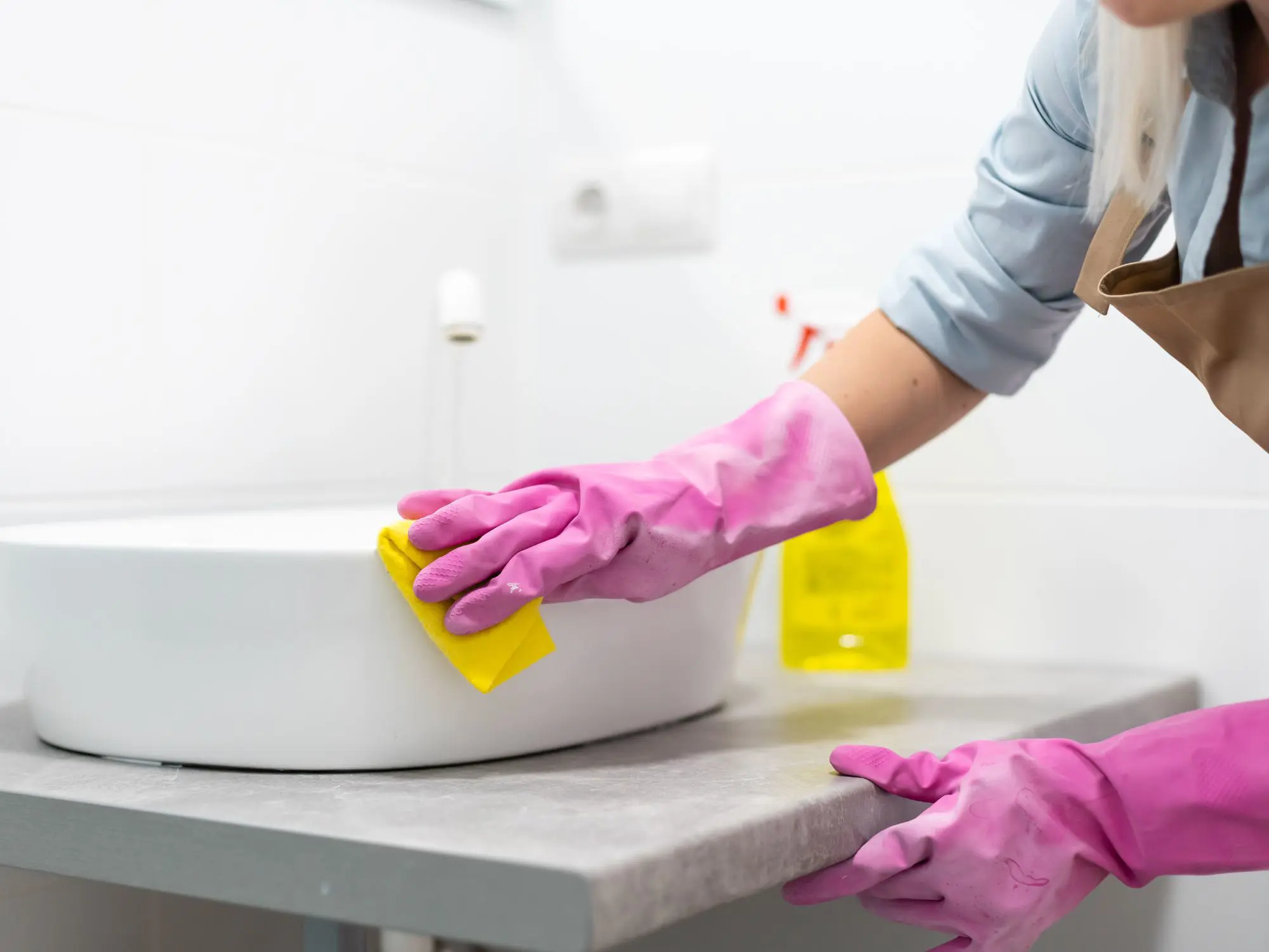 Person wearing pink rubber gloves cleaning a bathroom sink with a yellow sponge; a spray bottle with yellow liquid is visible in the background
