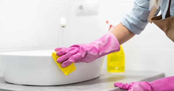 Person wearing pink rubber gloves cleaning a bathroom sink with a yellow sponge; a spray bottle with yellow liquid is visible in the background