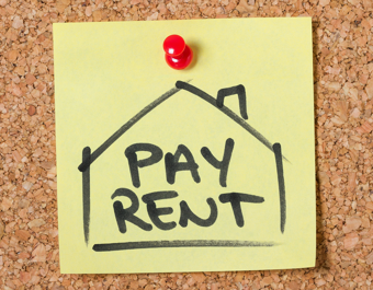 Yellow sticky note pinned to a corkboard with a red pushpin, featuring a drawing of a house and the words 'PAY RENT' written in bold black letters