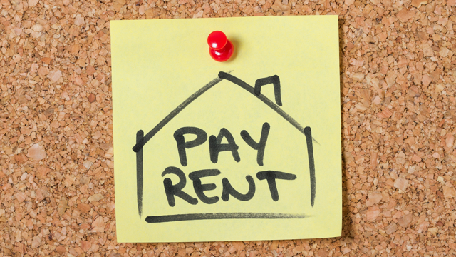 Yellow sticky note pinned to a corkboard with a red pushpin, featuring a drawing of a house and the words 'PAY RENT' written in bold black letters