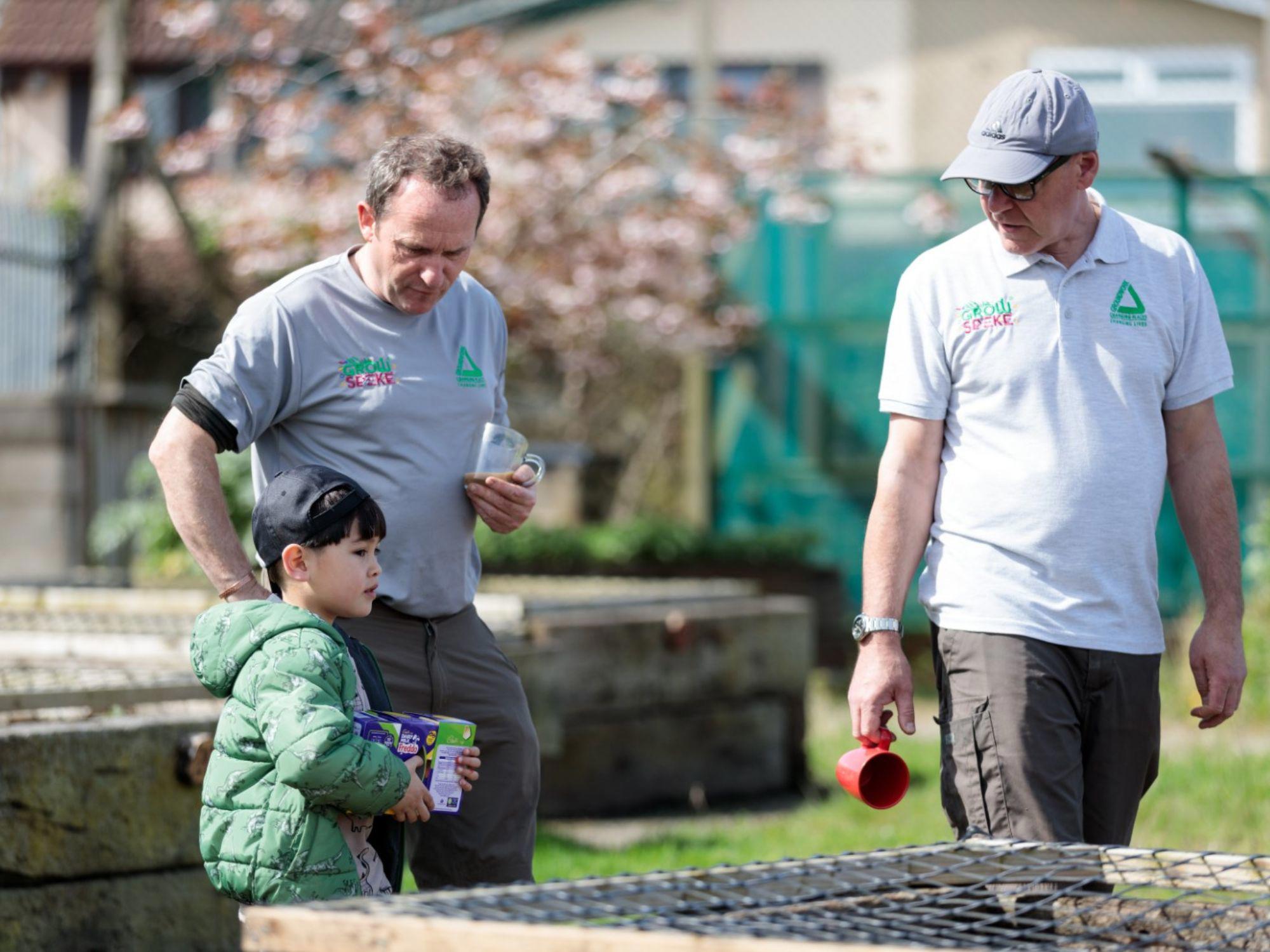 Two adults and a child in a garden setting with raised beds and greenhouse structures. The adults wear light-colored shirts with logos, one holding a red mug. The child wears a green jacket and holds a box of gardening supplies, suggesting an educational or community gardening activity.