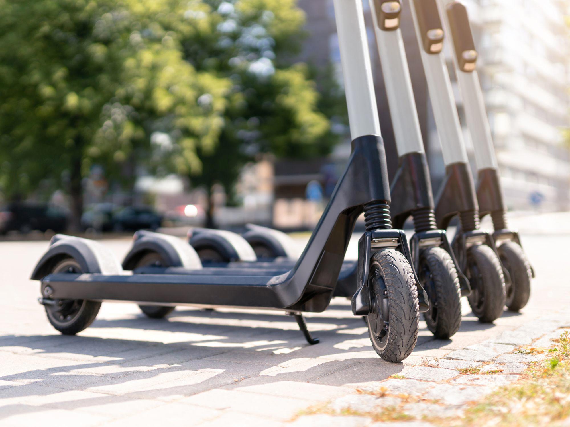 A row of electric scooters parked on a paved surface in an urban setting. The scooters are neatly lined up, showing handlebars and front wheels. Greenery and blurred buildings appear in the background.