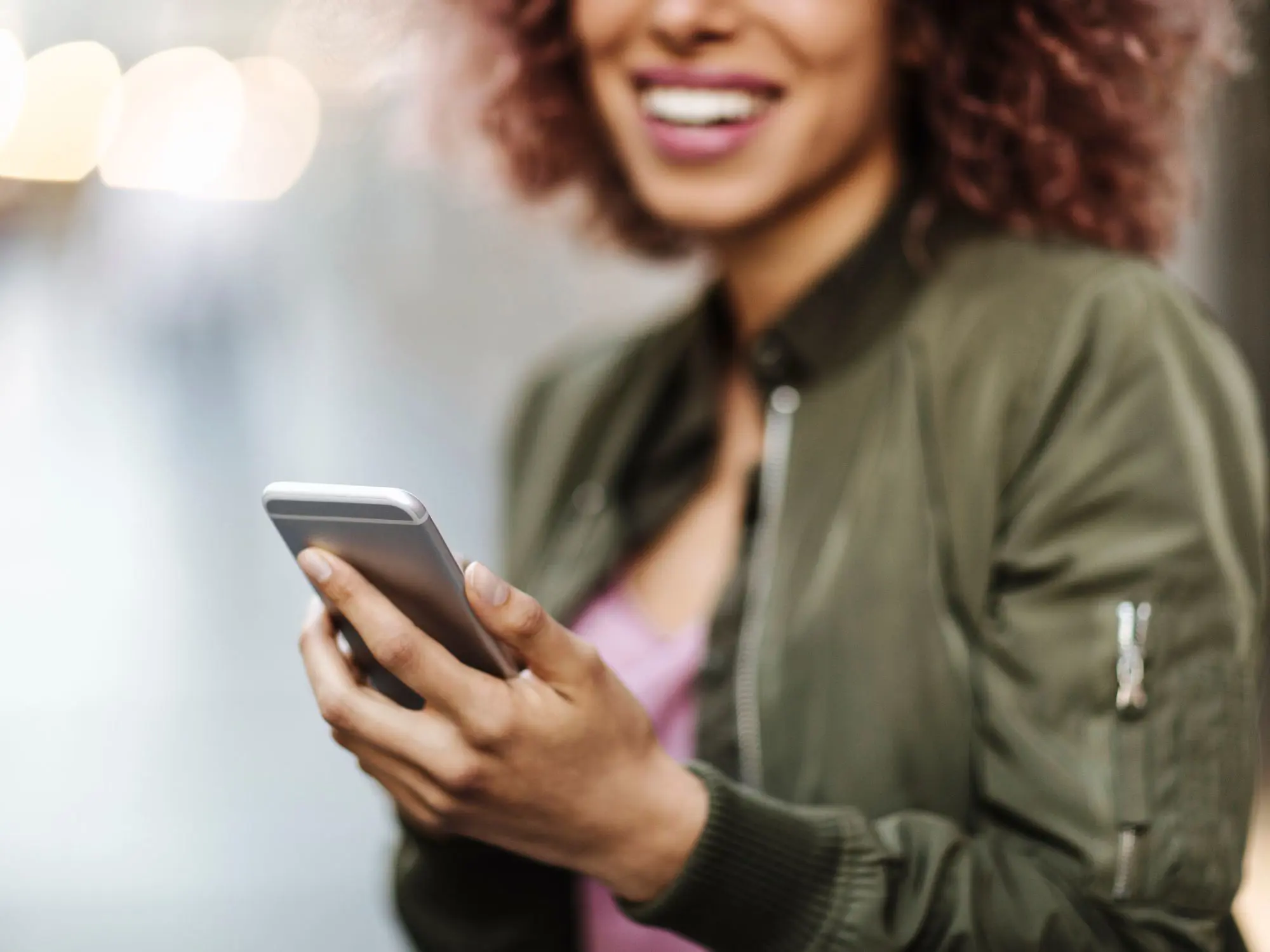 Person with curly hair wearing a green jacket, holding a smartphone with both hands in an outdoor setting with blurred lights in the background.