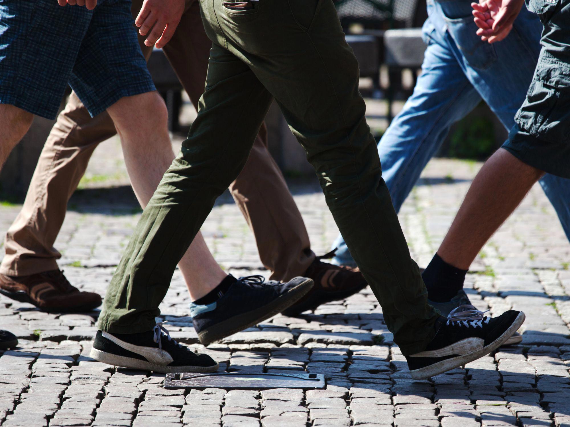 A group of people walking on a cobblestone street, shown from the knees down. Footwear includes sneakers, sandals, and casual shoes, with varied clothing styles like jeans, shorts, and khakis.