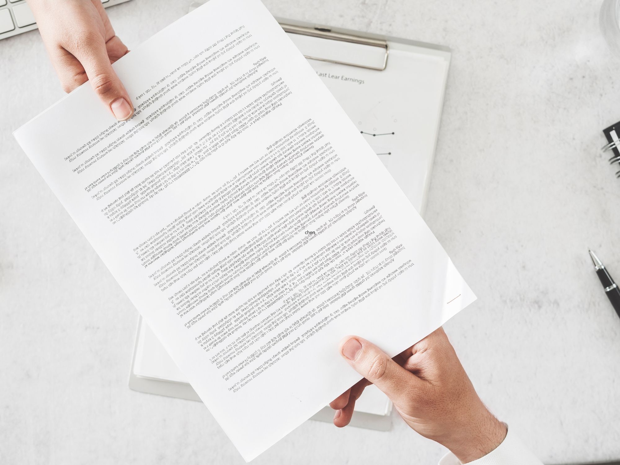 Two hands exchange a printed document over a clipboard with papers and a pen in the background; the document contains multiple paragraphs of text, though the content is not legible.