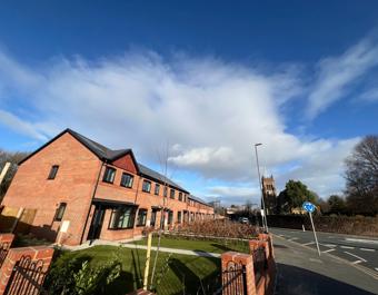 A row of newly built red‑brick terraced houses with black window frames sits on a corner plot under a bright blue sky with scattered clouds. A neat lawn and young trees line the front, bordered by low brick walls and metal railings. A road with a mini-roundabout runs alongside, and a large church tower is visible in the background among mature trees.