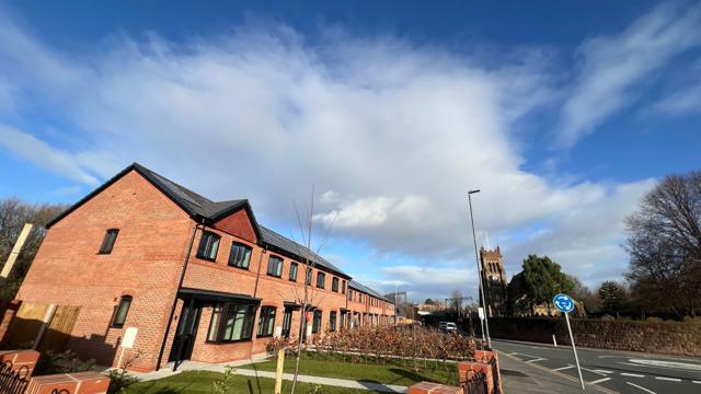 A row of newly built red‑brick terraced houses with black window frames sits on a corner plot under a bright blue sky with scattered clouds. A neat lawn and young trees line the front, bordered by low brick walls and metal railings. A road with a mini-roundabout runs alongside, and a large church tower is visible in the background among mature trees.