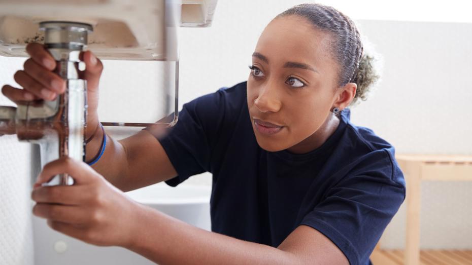 Person working under a sink, adjusting plumbing pipes with both hands; a toilet and wooden shelf are visible in the background. Person working under a sink, adjusting plumbing pipes with both hands; a toilet and wooden shelf are visible in the background.
