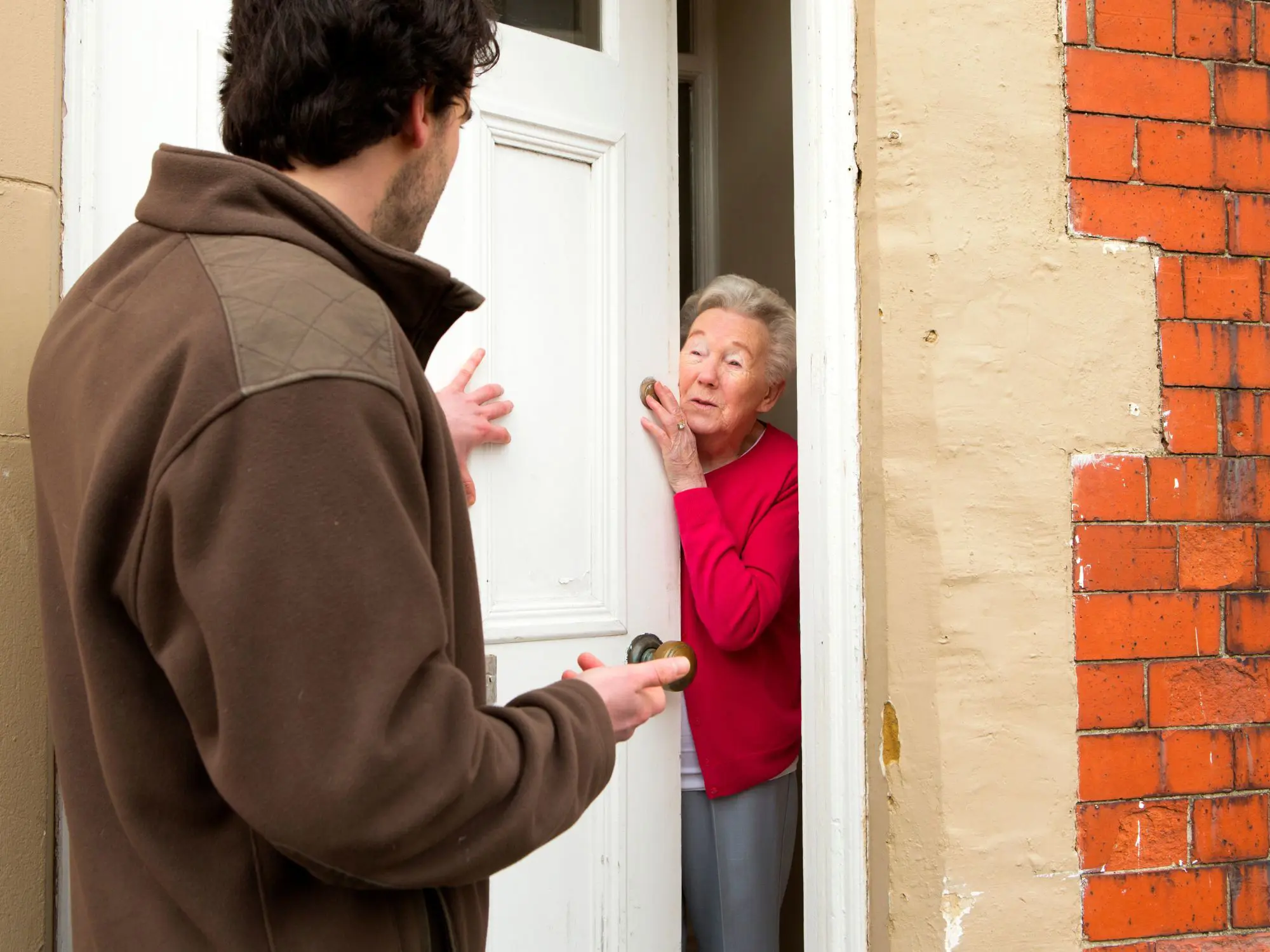 A person in a brown jacket stands at the doorway of a house, speaking to someone inside. The door is white, and the exterior wall features red bricks and beige plaster.