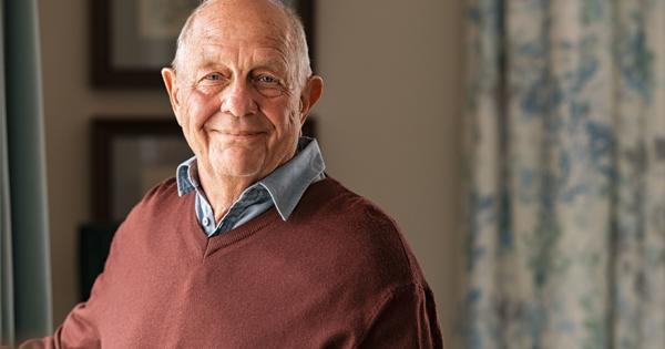 Person wearing a maroon sweater over a blue collared shirt, with framed pictures and patterned curtains in the background.