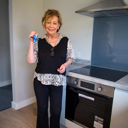 A person stands in a newly fitted kitchen holding a set of keys. The kitchen features light-coloured units, a built-in oven, induction hob, stainless-steel extractor hood and grey splashback, with wood-effect flooring throughout.