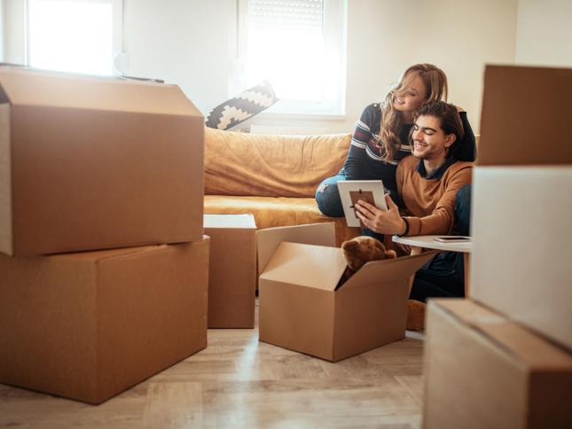 Two people sitting on the floor among several cardboard boxes, unpacking belongings in a bright living room with a mustard-coloured sofa. Two people sitting on the floor among several cardboard boxes, unpacking belongings in a bright living room with a mustard-coloured sofa.