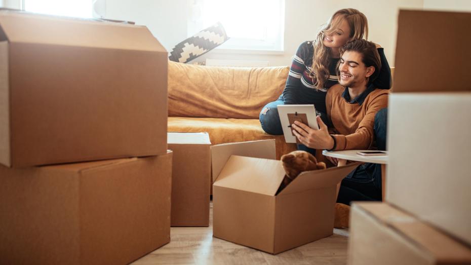 Two people sitting on the floor among several cardboard boxes, unpacking belongings in a bright living room with a mustard-coloured sofa. Two people sitting on the floor among several cardboard boxes, unpacking belongings in a bright living room with a mustard-coloured sofa.