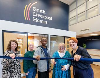 Five people stand in front of a building with a “South Liverpool Homes” sign, holding a blue ribbon in a ribbon-cutting ceremony. Behind them are glass doors and windows, with the slogan “Great homes | Strong communities | Bright futures” visible on the wall.