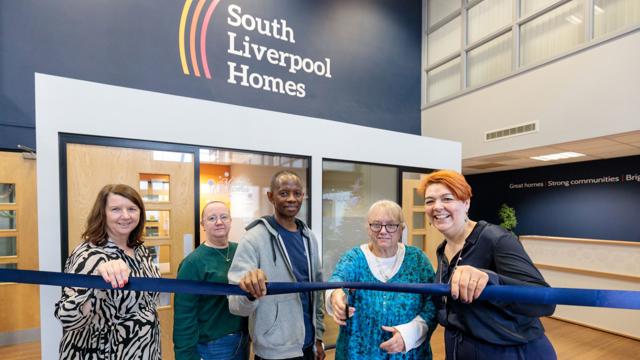 Five people stand in front of a building with a “South Liverpool Homes” sign, holding a blue ribbon in a ribbon-cutting ceremony. Behind them are glass doors and windows, with the slogan “Great homes | Strong communities | Bright futures” visible on the wall.