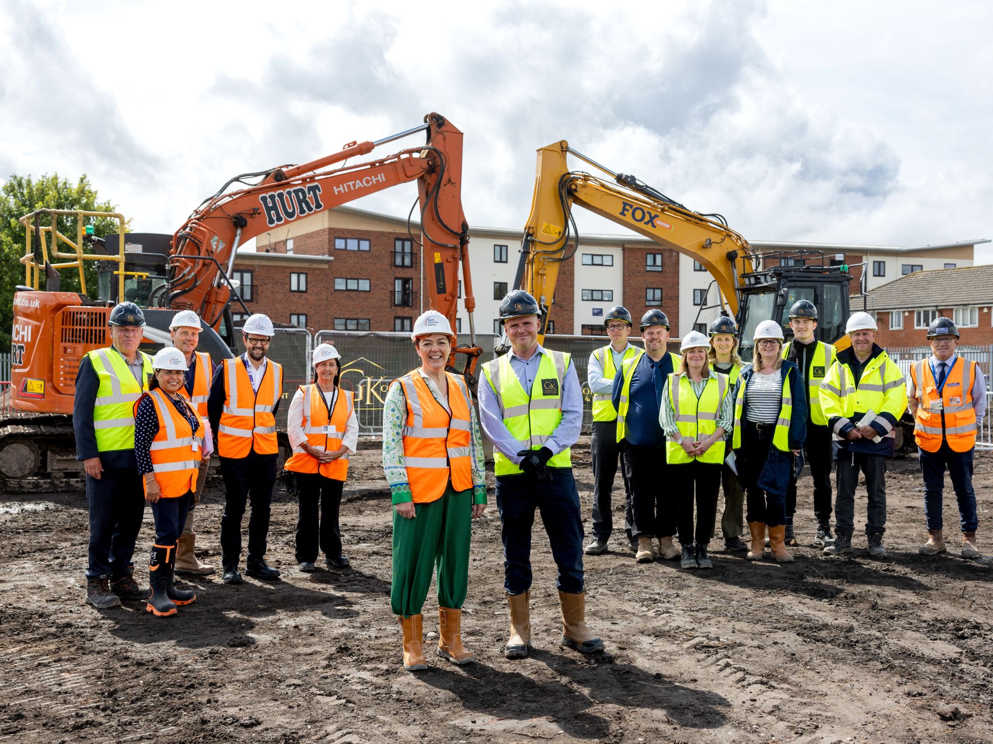 Group of people in high-visibility vests and hard hats standing on an urban construction site, with two excavators in the background.