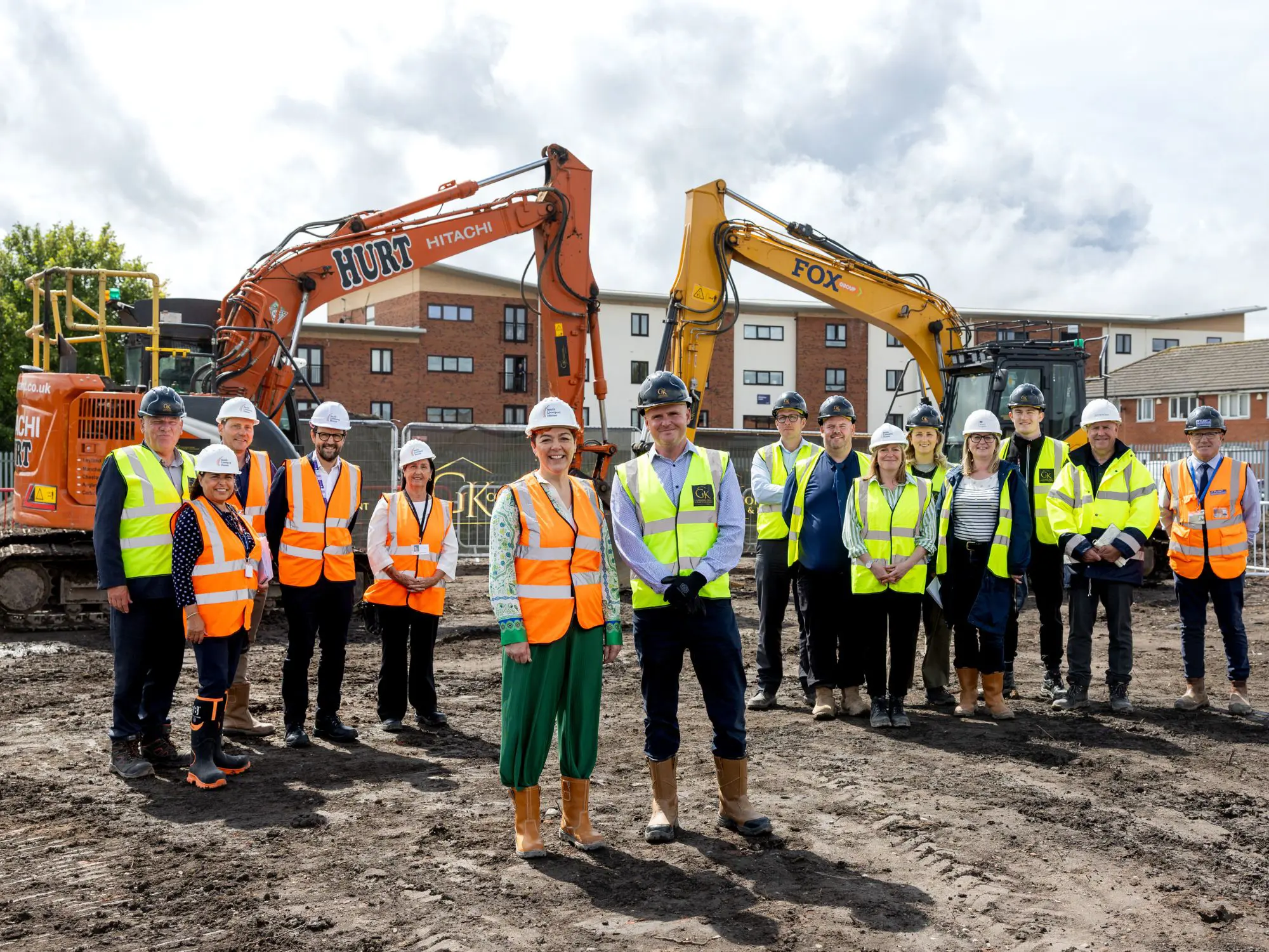 Group of people in high-visibility vests and hard hats standing on an urban construction site, with two excavators in the background.