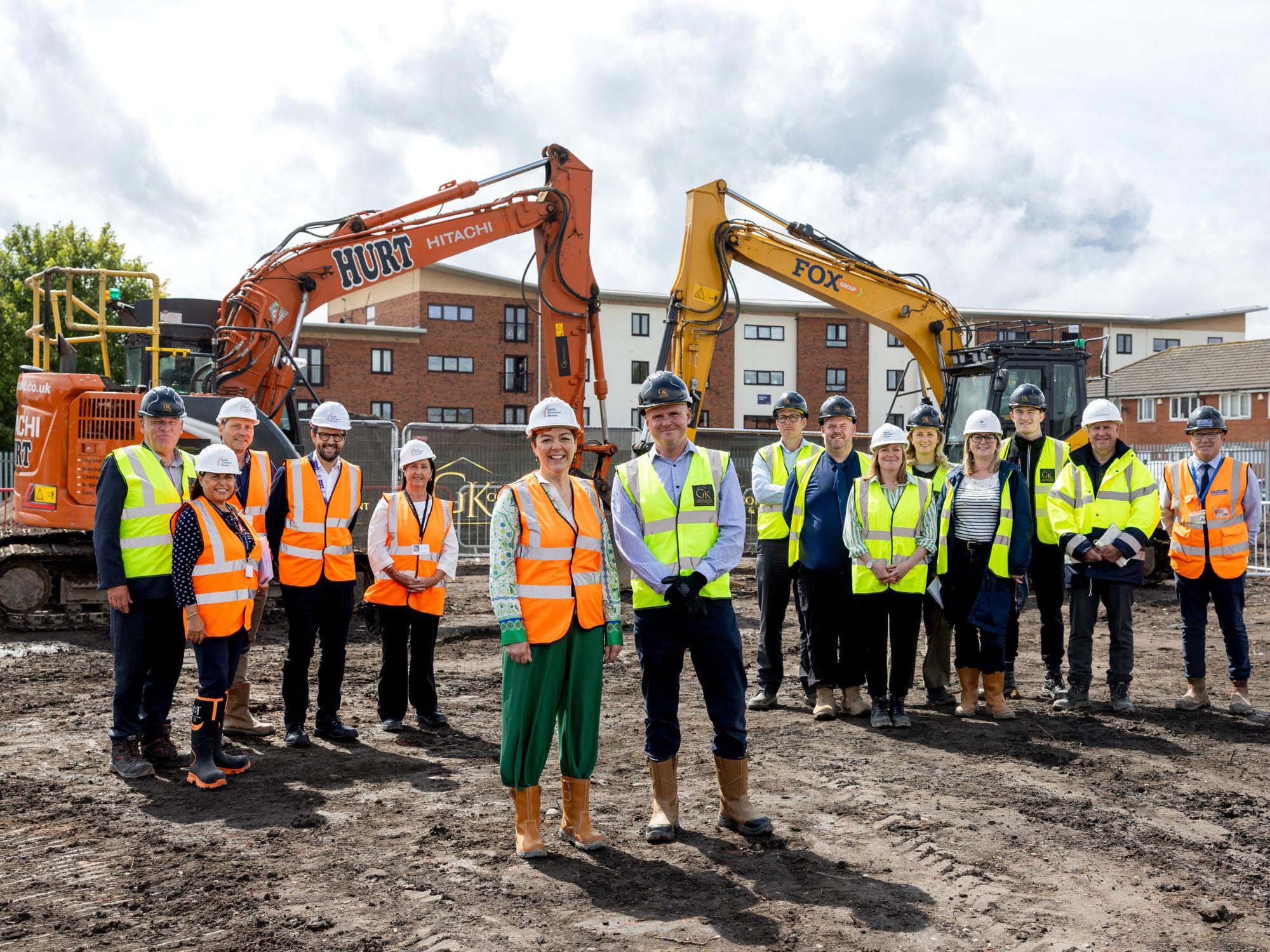 Group of people in high-visibility vests and hard hats standing on an urban construction site, with two excavators in the background.