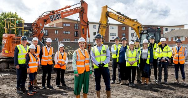 Group of people in high-visibility vests and hard hats standing on an urban construction site, with two excavators in the background.