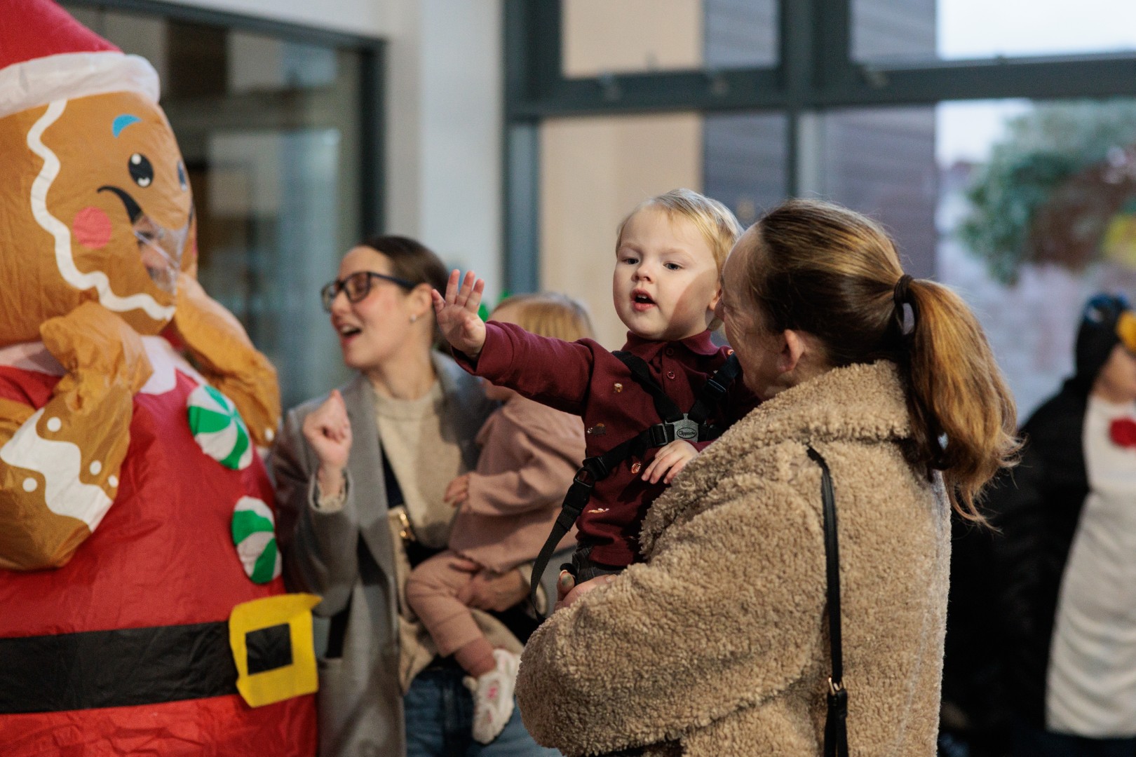 A parent holds a young child who is reaching towards a person in a colourful inflatable gingerbread costume during a festive indoor event. Other families stand nearby enjoying the activity.