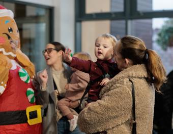 A parent holds a young child who is reaching towards a person in a colourful inflatable gingerbread costume during a festive indoor event. Other families stand nearby enjoying the activity.