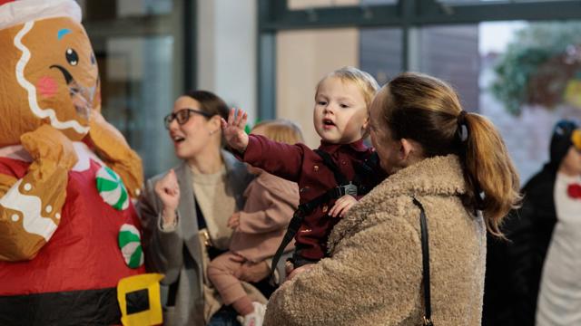 A parent holds a young child who is reaching towards a person in a colourful inflatable gingerbread costume during a festive indoor event. Other families stand nearby enjoying the activity.