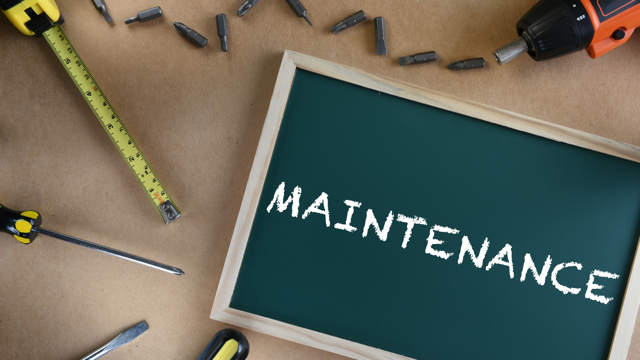 A small chalkboard with the word 'MAINTENANCE' written in white chalk, surrounded by tools including a tape measure, screwdriver, drill bits, and a power drill, representing repair or upkeep work.
