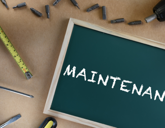 A small chalkboard with the word 'MAINTENANCE' written in white chalk, surrounded by tools including a tape measure, screwdriver, drill bits, and a power drill, representing repair or upkeep work.