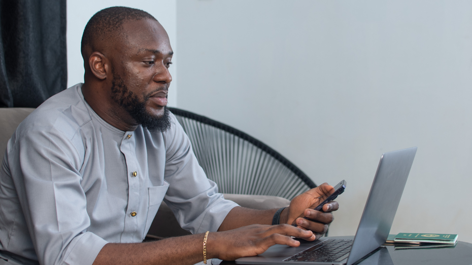 A person sitting at a small table using a laptop while holding a smartphone, with a passport and documents placed beside the laptop.