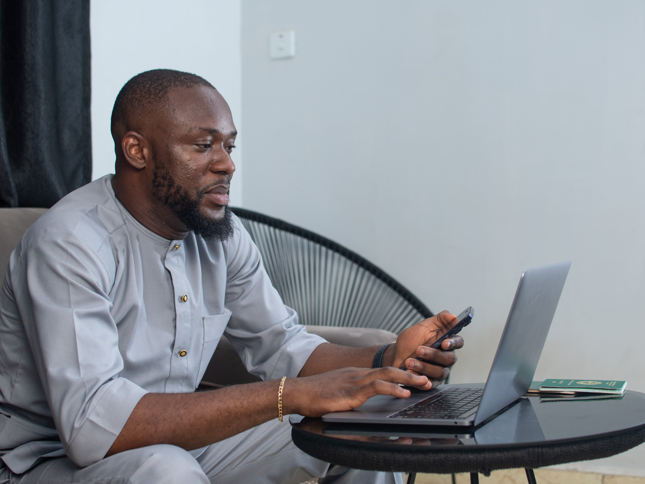 A person sitting at a small table using a laptop while holding a smartphone, with a passport and documents placed beside the laptop.