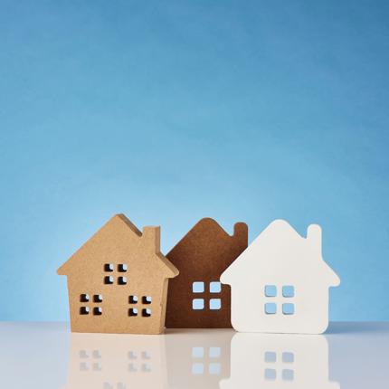 Three house-shaped cutouts with four square windows each, made of brown, dark brown, and white materials, placed side by side on a reflective surface against a blue background.