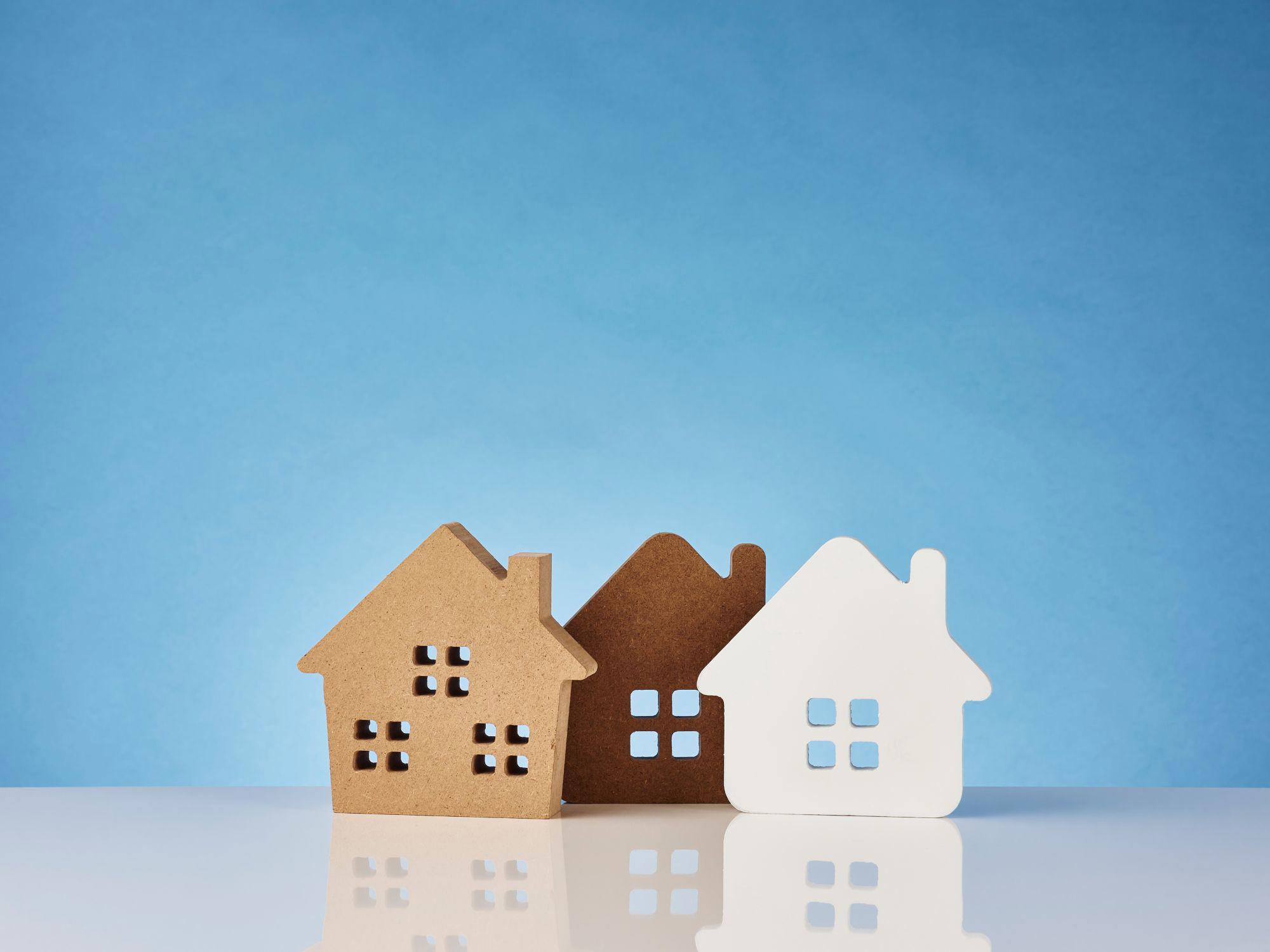 Three house-shaped cutouts with four square windows each, made of brown, dark brown, and white materials, placed side by side on a reflective surface against a blue background.