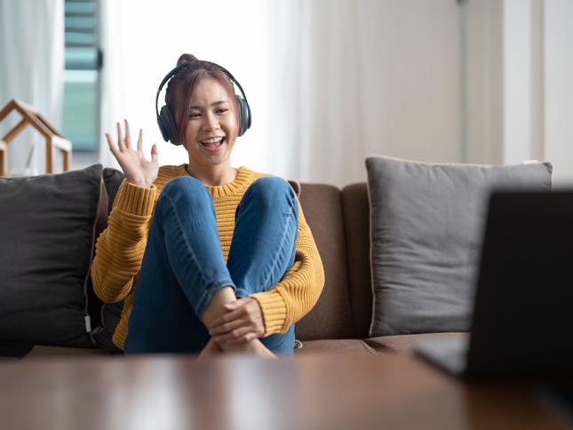 A person wearing headphones sitting on a couch, waving at a laptop screen during a video call. A person wearing headphones sitting on a couch, waving at a laptop screen during a video call.