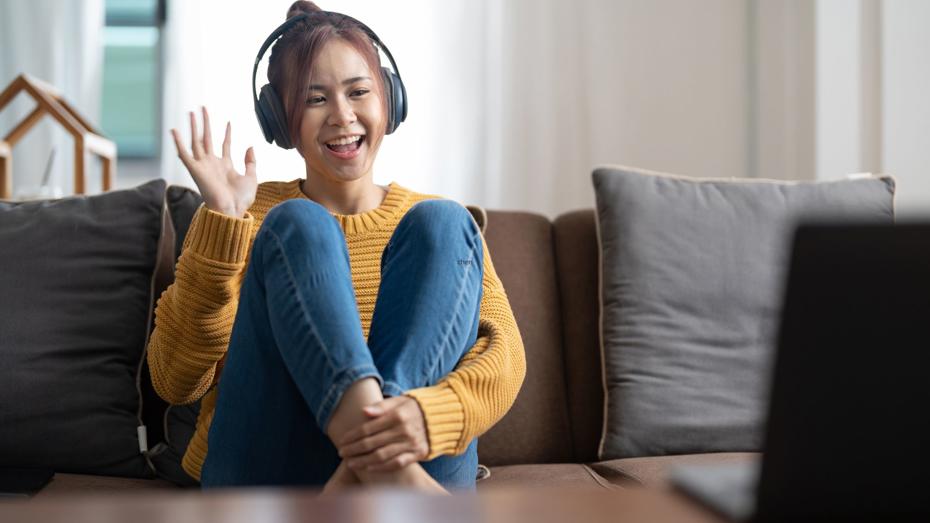 A person wearing headphones sitting on a couch, waving at a laptop screen during a video call. A person wearing headphones sitting on a couch, waving at a laptop screen during a video call.