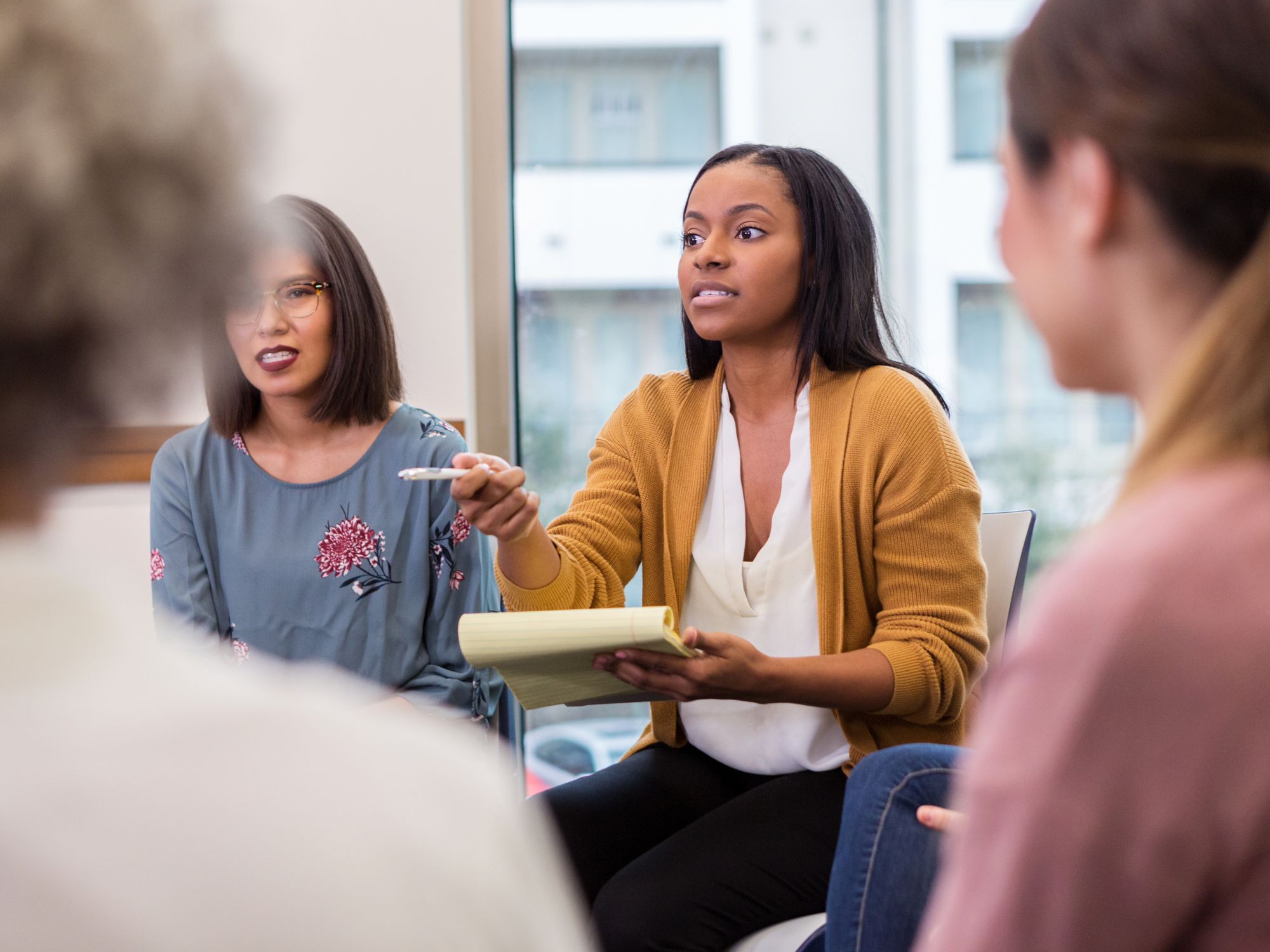 "A group of people in a meeting room engaged in discussion. One person is holding a notepad and pen, actively participating. Large windows are visible in the background, suggesting an indoor office setting."