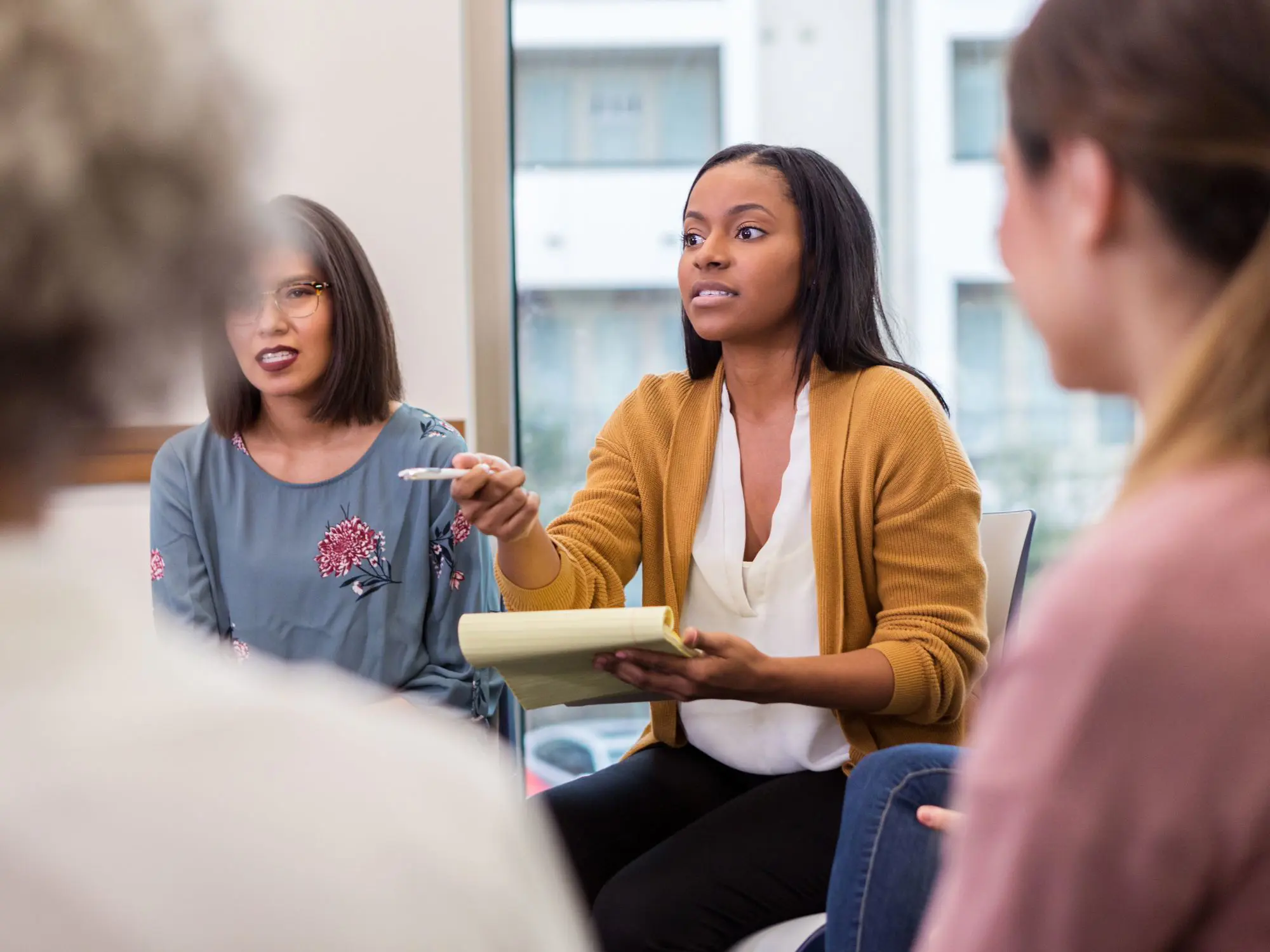 "A group of people in a meeting room engaged in discussion. One person is holding a notepad and pen, actively participating. Large windows are visible in the background, suggesting an indoor office setting."