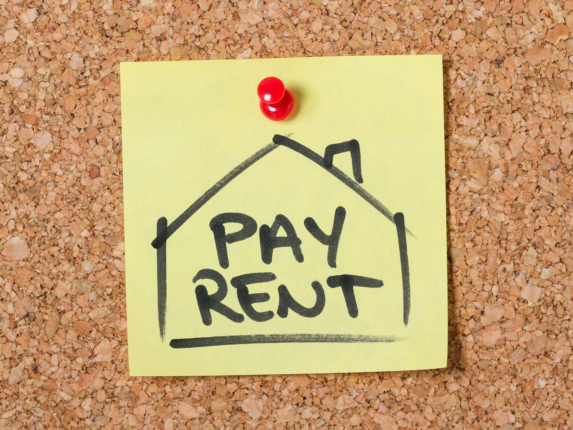 A yellow sticky note pinned to a corkboard with a red pushpin, featuring a drawing of a house and the bold text 'PAY RENT' inside—serving as a visual reminder to pay rent.