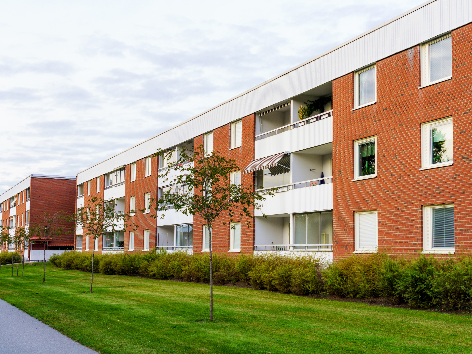 Modern three-story apartment building with red brick and white paneling, featuring balconies and a landscaped lawn with trees and shrubs.