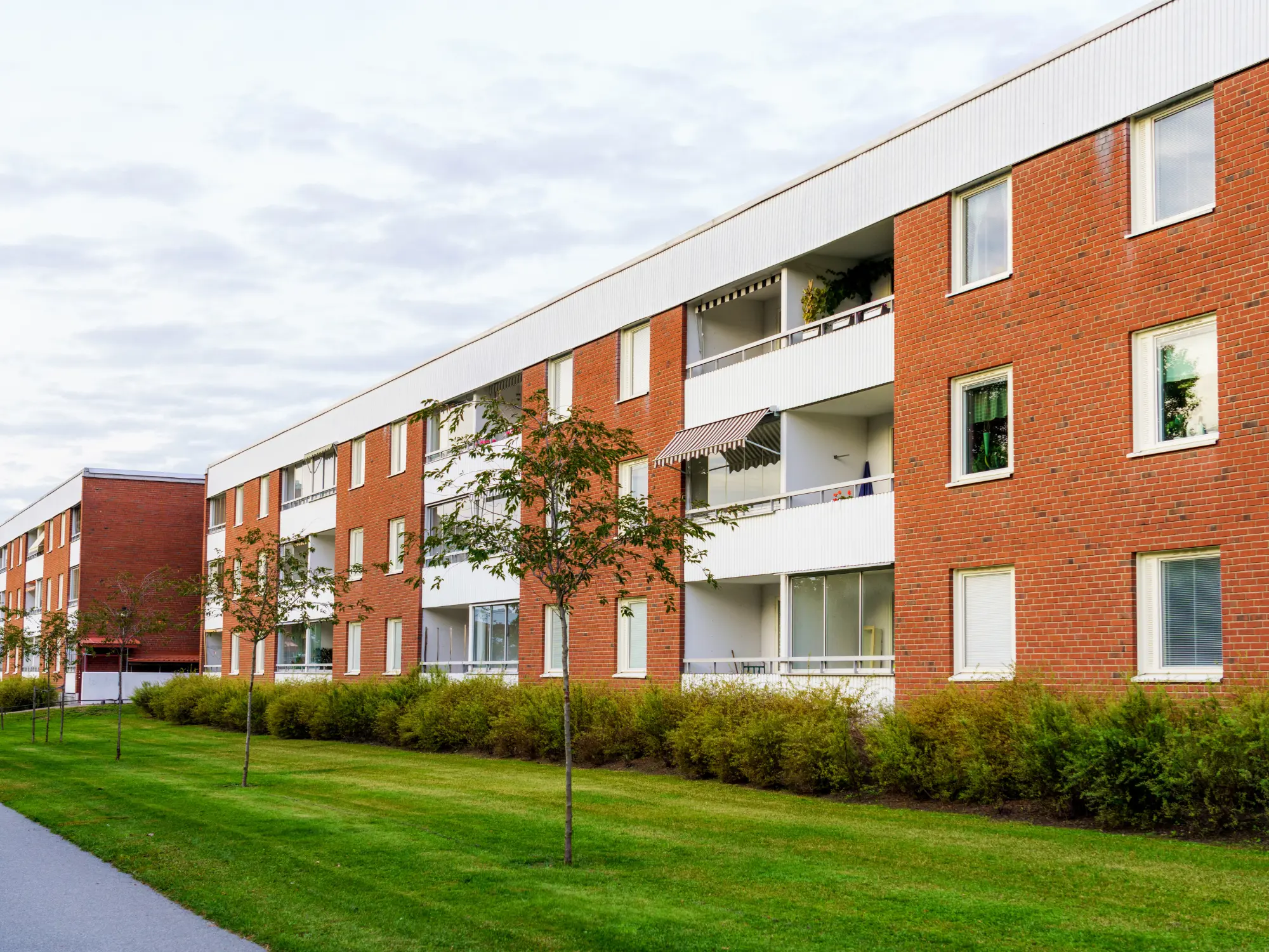 Modern three-story apartment building with red brick and white paneling, featuring balconies and a landscaped lawn with trees and shrubs.